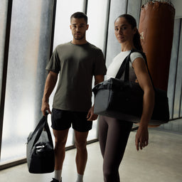 A man and woman standing in a gym with a boxing bag holding a gym duffel 25L in All Black
