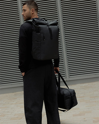 A man standing in front of a wall with a black roll top 20l on his back and holding a gym duffel 25l in black by his side