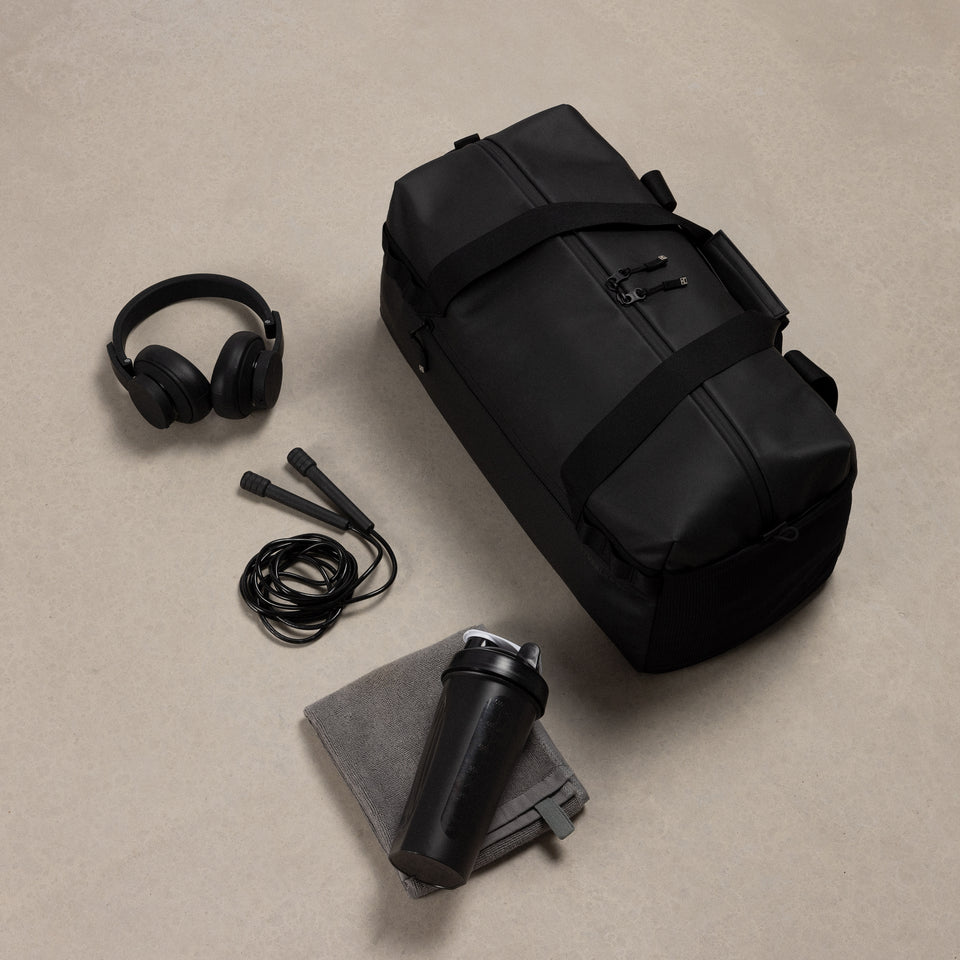 A studio shot of the gym duffel 25l in All Black with the content around including a skipping rope, headphones and towel