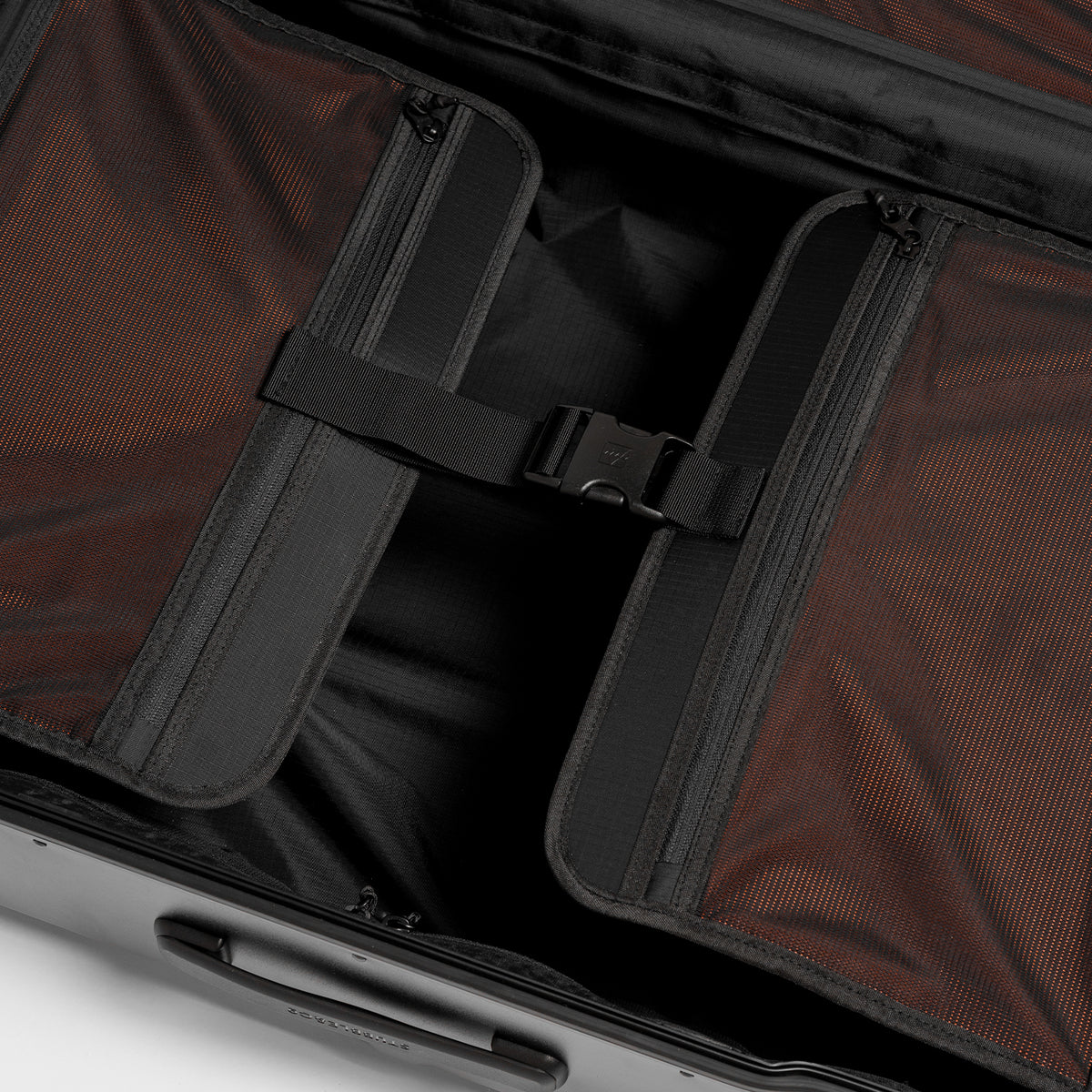 Close-up of the interior of a hard-shell suitcase showing a black zipper luggage organizer pocket with a strap and buckle, set against orange textured lining.