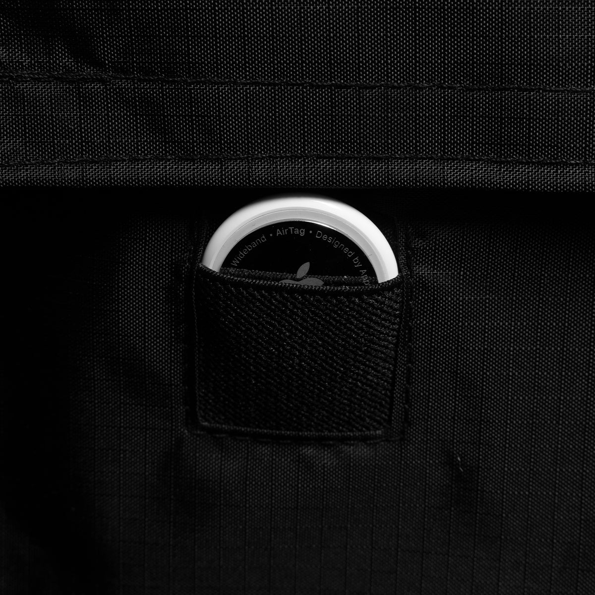 A close-up of a black textured fabric bag with a small front pocket holding a round white AirTag, photographed against a dark black background.