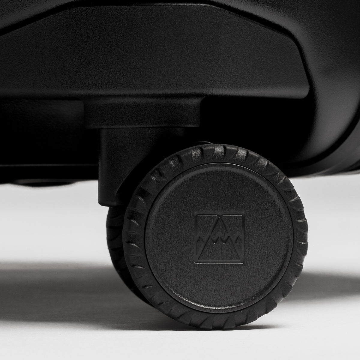 Close-up of a black wheeled suitcase or rolling bag, showing a textured black wheel with a square mountain-like logo embossed in the center, photographed on a light gray studio background.