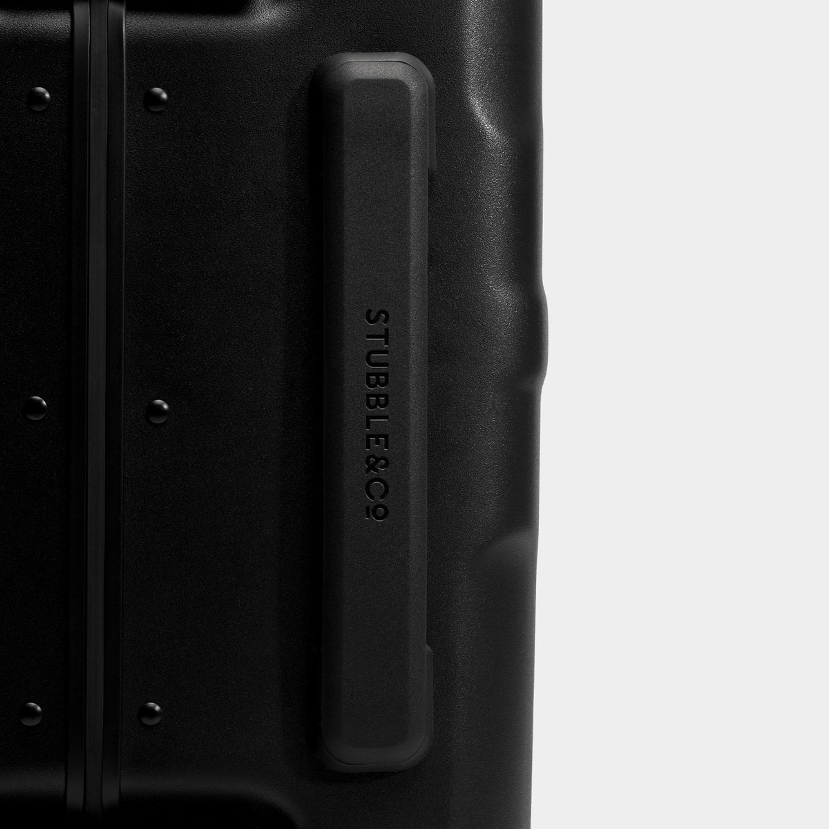 Close-up of a black hard-shell suitcase corner showing a recessed handle with the debossed brand name STUBBLE&Co on a light gray background.