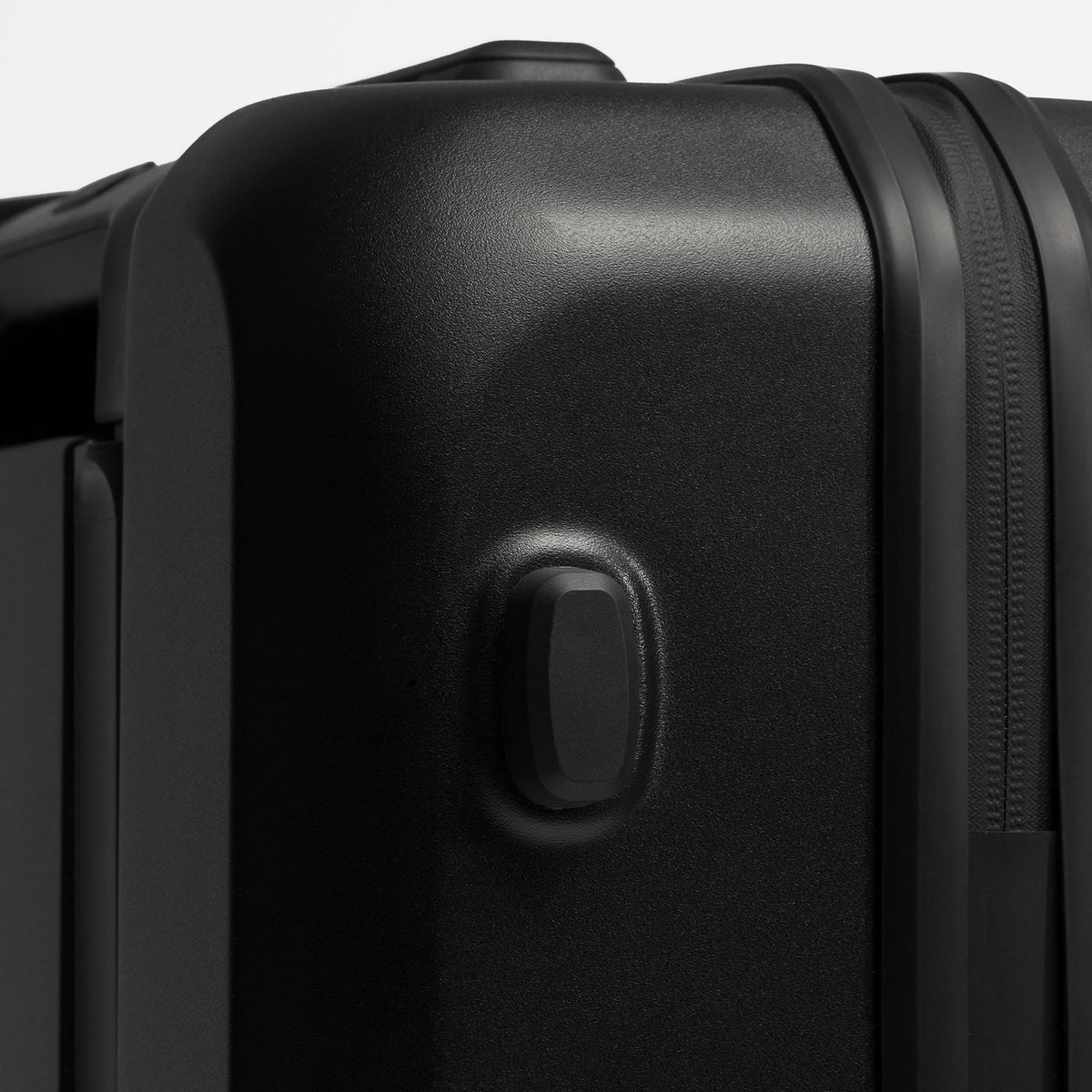 Close-up of a black hard-shell suitcase corner showing a recessed lock button and zipper track against a light gray background.