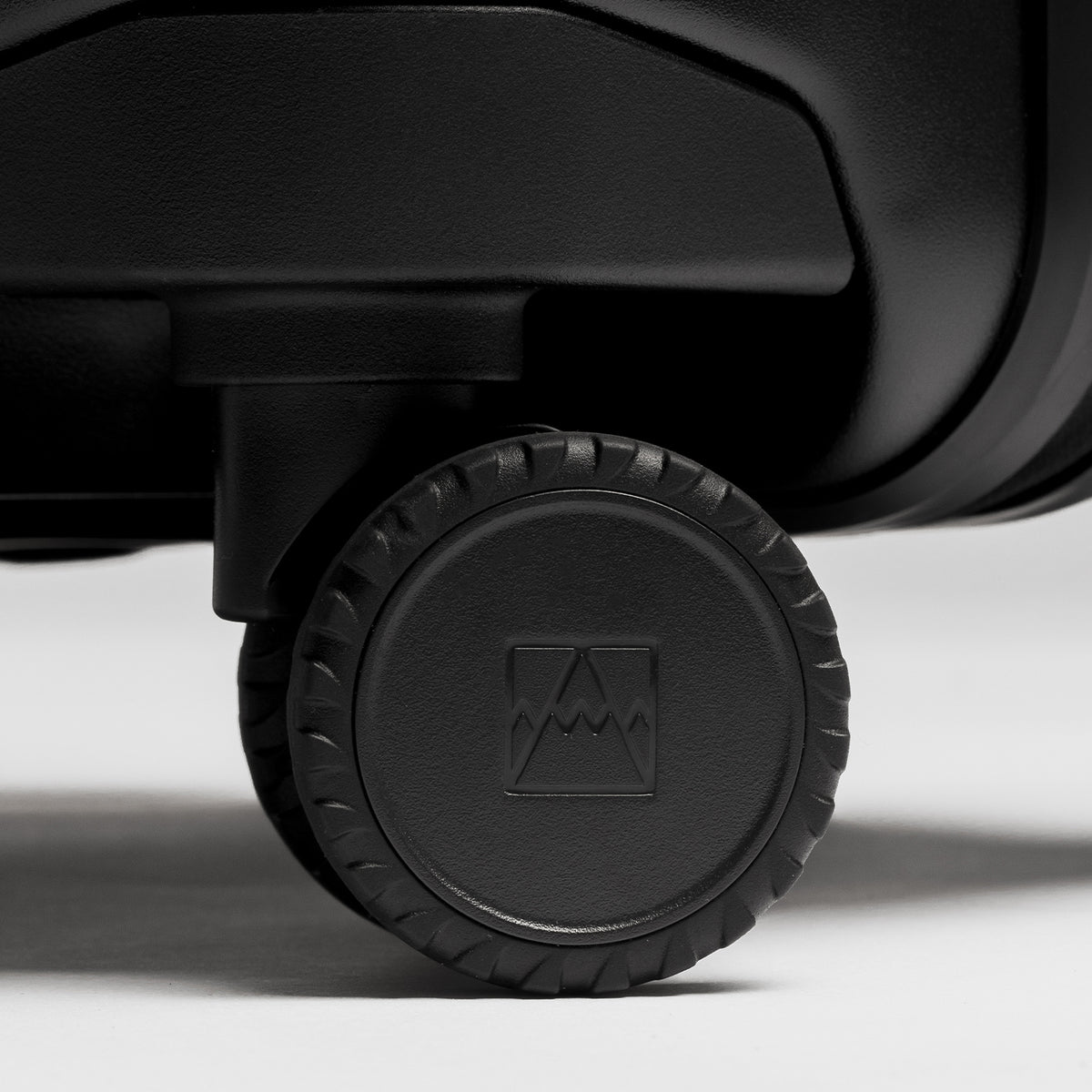 Close-up of a black hard-shell suitcase wheel with a textured tread and an embossed mountain logo in the center, set against a clean light gray background.