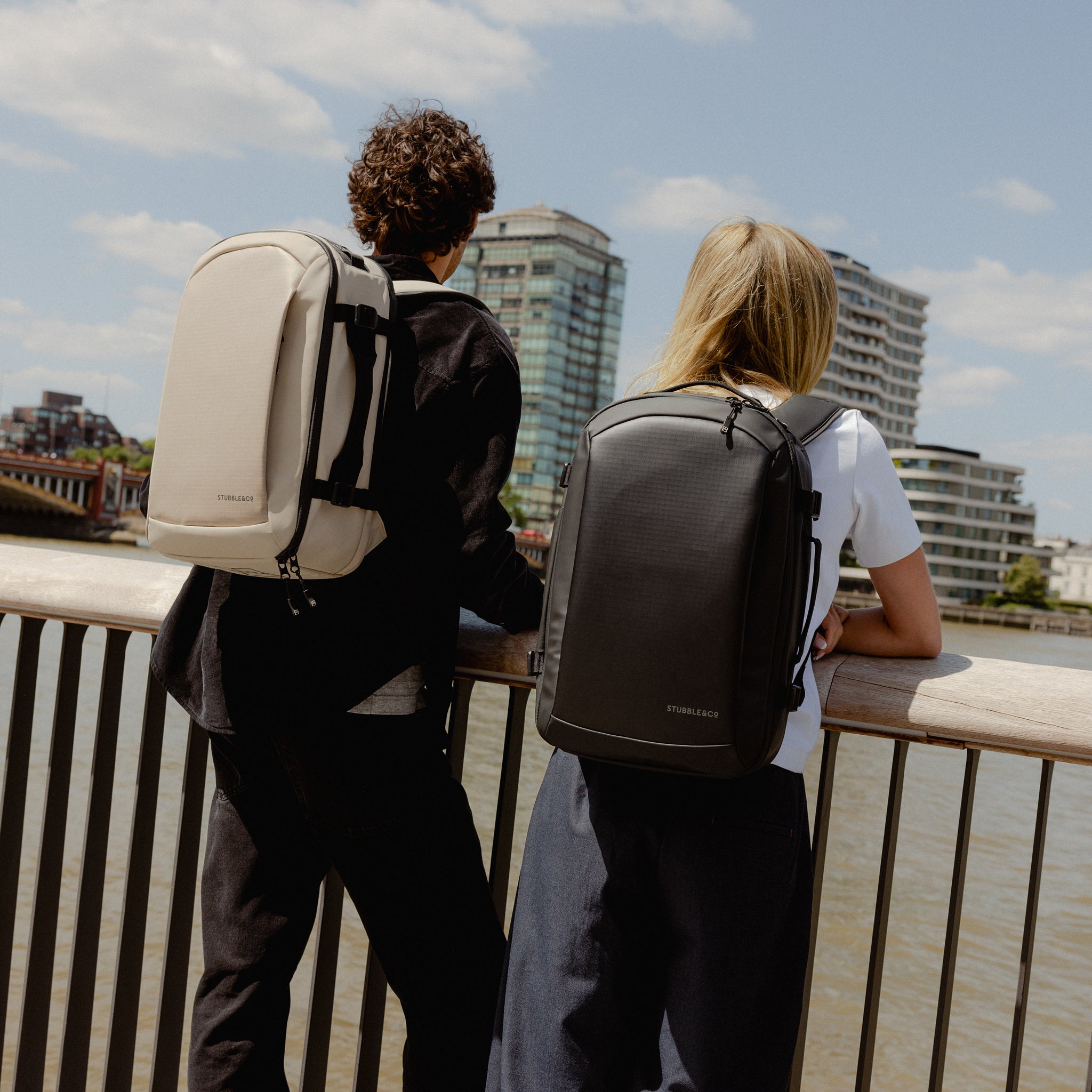 A man and woman standing by the river wearing Hybrid backpack 20L on their backs in All Black and sand