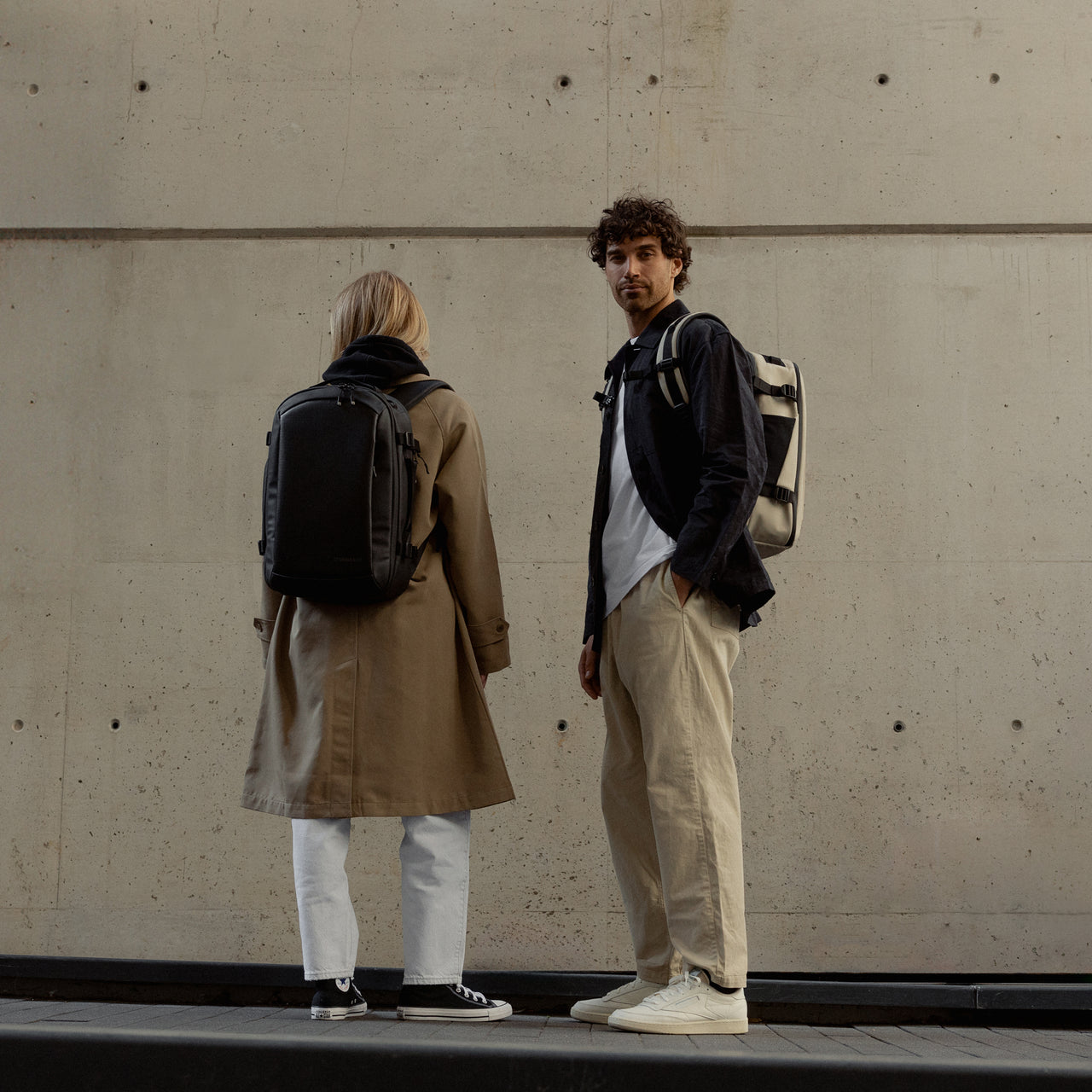 A man and woman wearing Hybrid Backpack 20L in sand and All Black on their backs