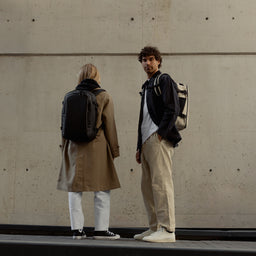 A man and woman wearing Hybrid Backpack 20L in sand and All Black on their backs