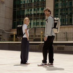 A man and woman standing side on wearing Hybrid Backpack 20L on their backs in All Black and sand