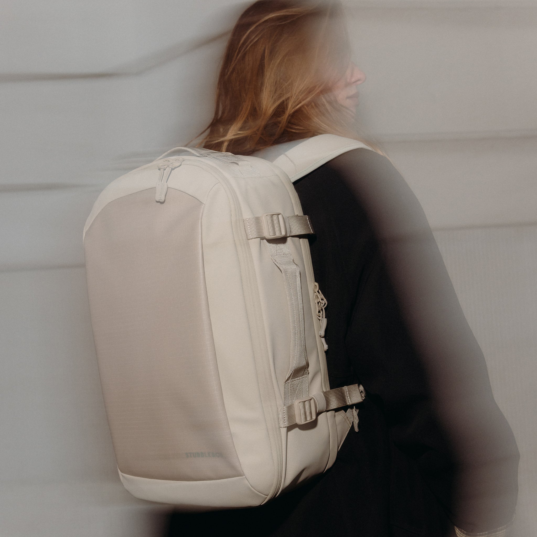 A woman wearing the Tonal Sand Hybrid Backpack 20L on her back with a blurred affect but the back in focus
