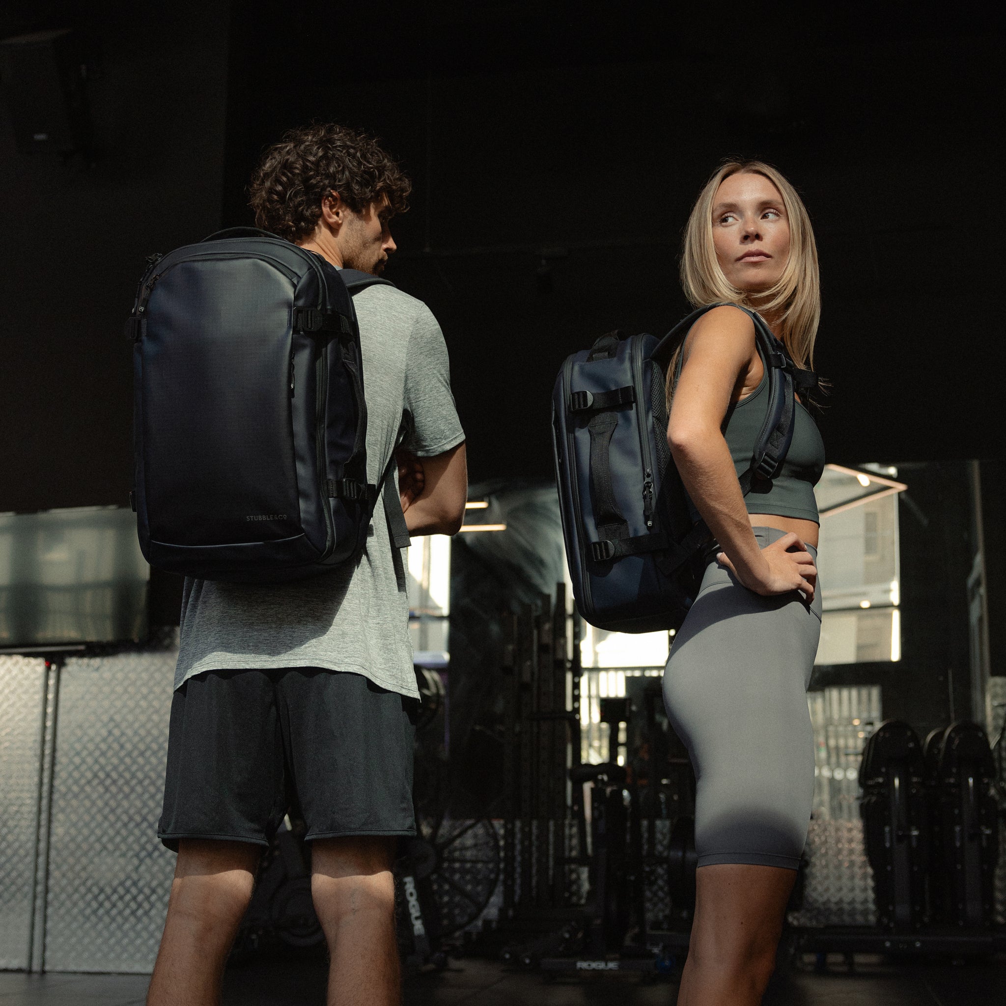 A man and woman standing in a gym and they are both wearing Hybrid Backpacks in Midnight Blue on her back