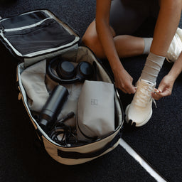 A woman tying up her shoes next to her open Hybrid backpack 30L in Sand packed with gym equipment