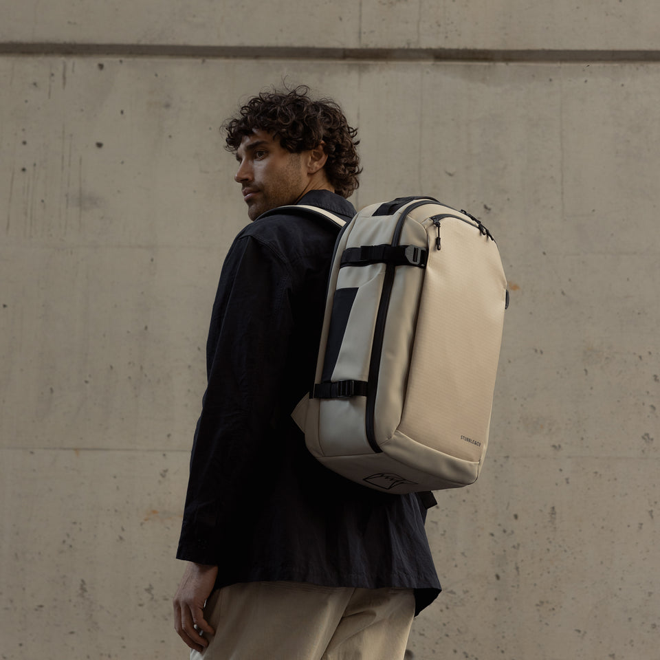 A man standing side on wearing a Hybrid Backpack 30L in sand on his back