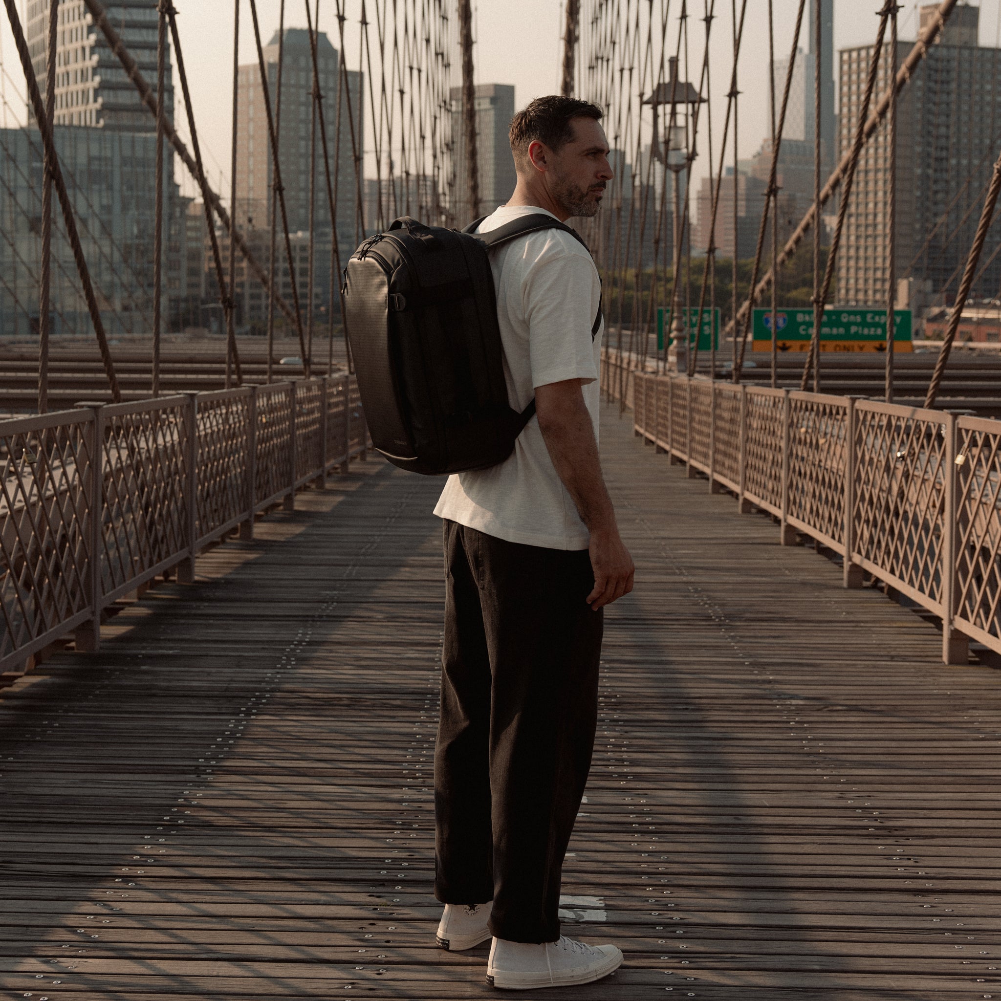 A lifestyle shot of a man standing on Brooklyn Bridge in New york with a Hybrid Backpack 30L in All Black on his back