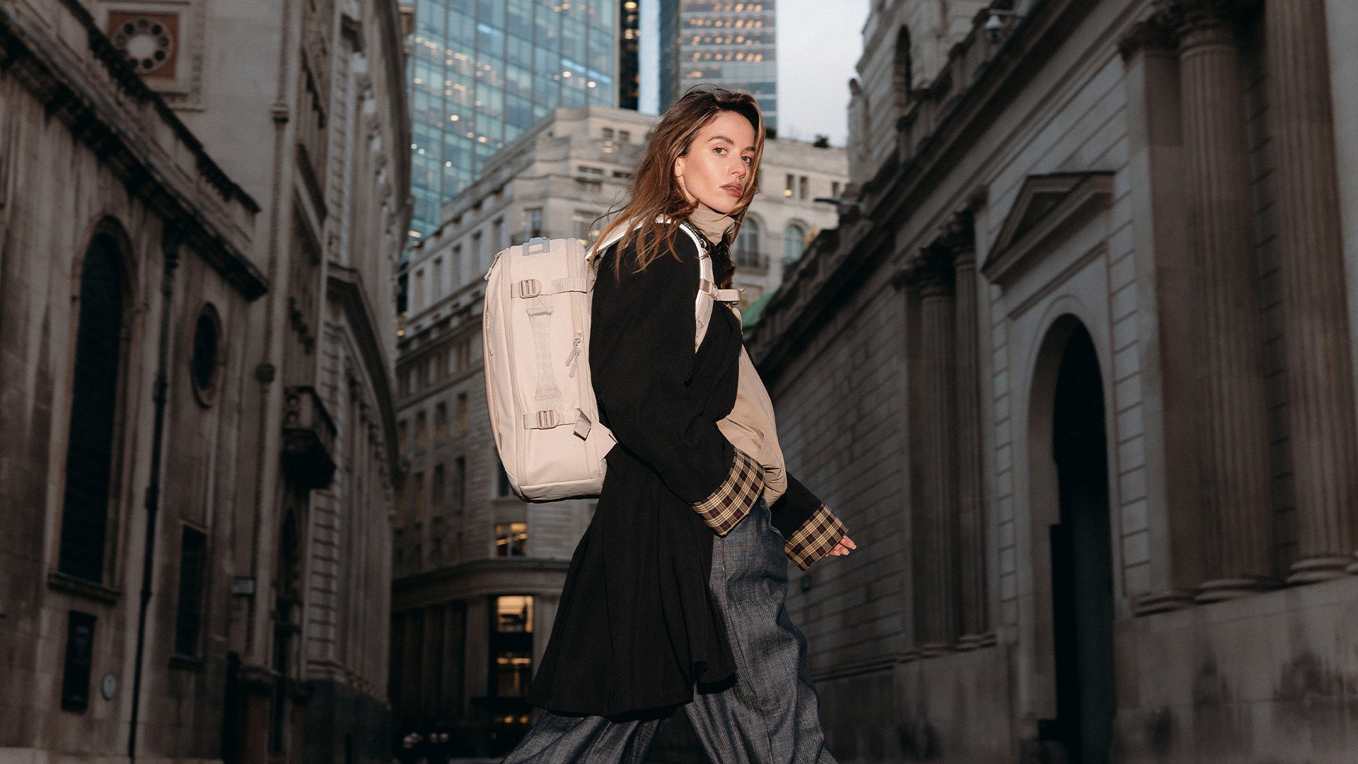 A woman crossing the street in London wearing a hybrid backpacks 20l in tonal sand on her back as she looks at the camera