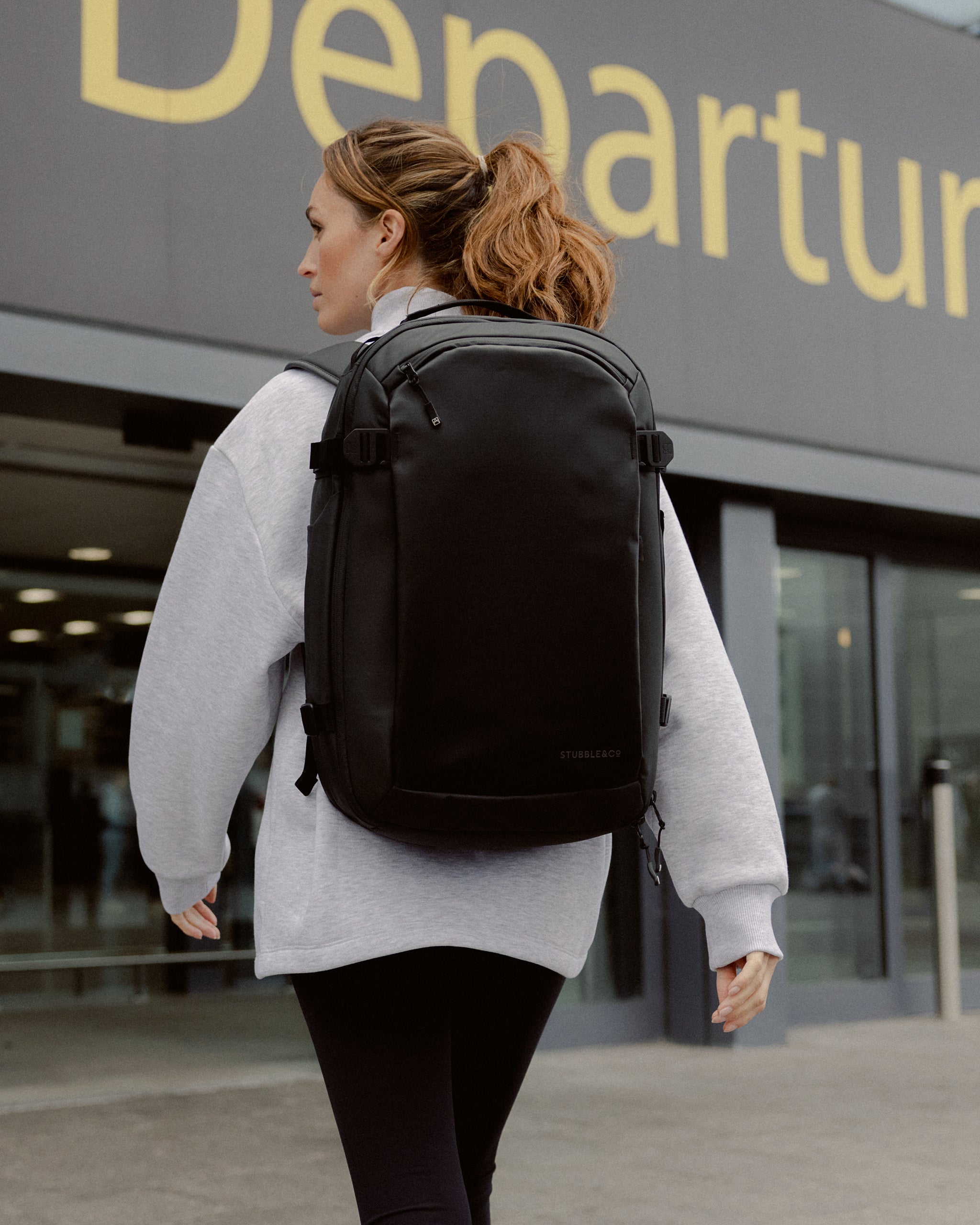 A woman walking into an airport departure lounge wearing a black Hybrid Backpack 30L on her back