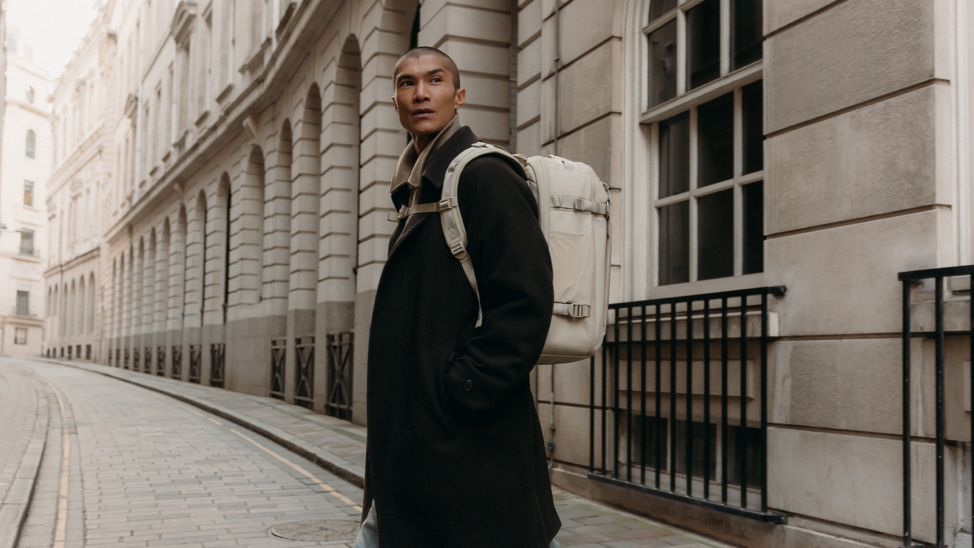 A man walking across the street wearing a Tonal Sand Hybrid Backpack 30L on his back with the chest straps done up