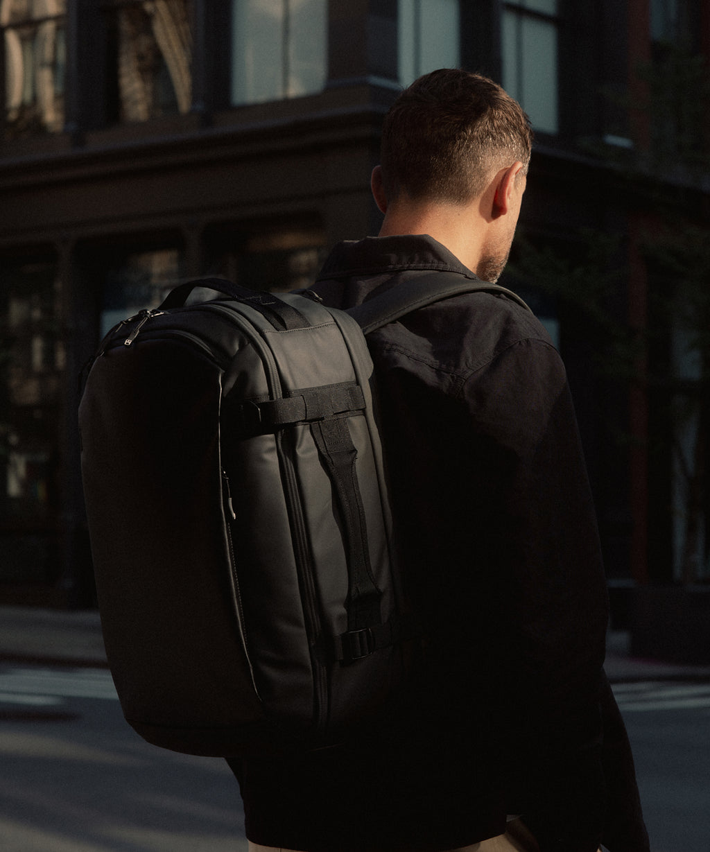 A man viewed from behind wearing a black backpack with a large front slip pocket, standing on a city street at dusk.
