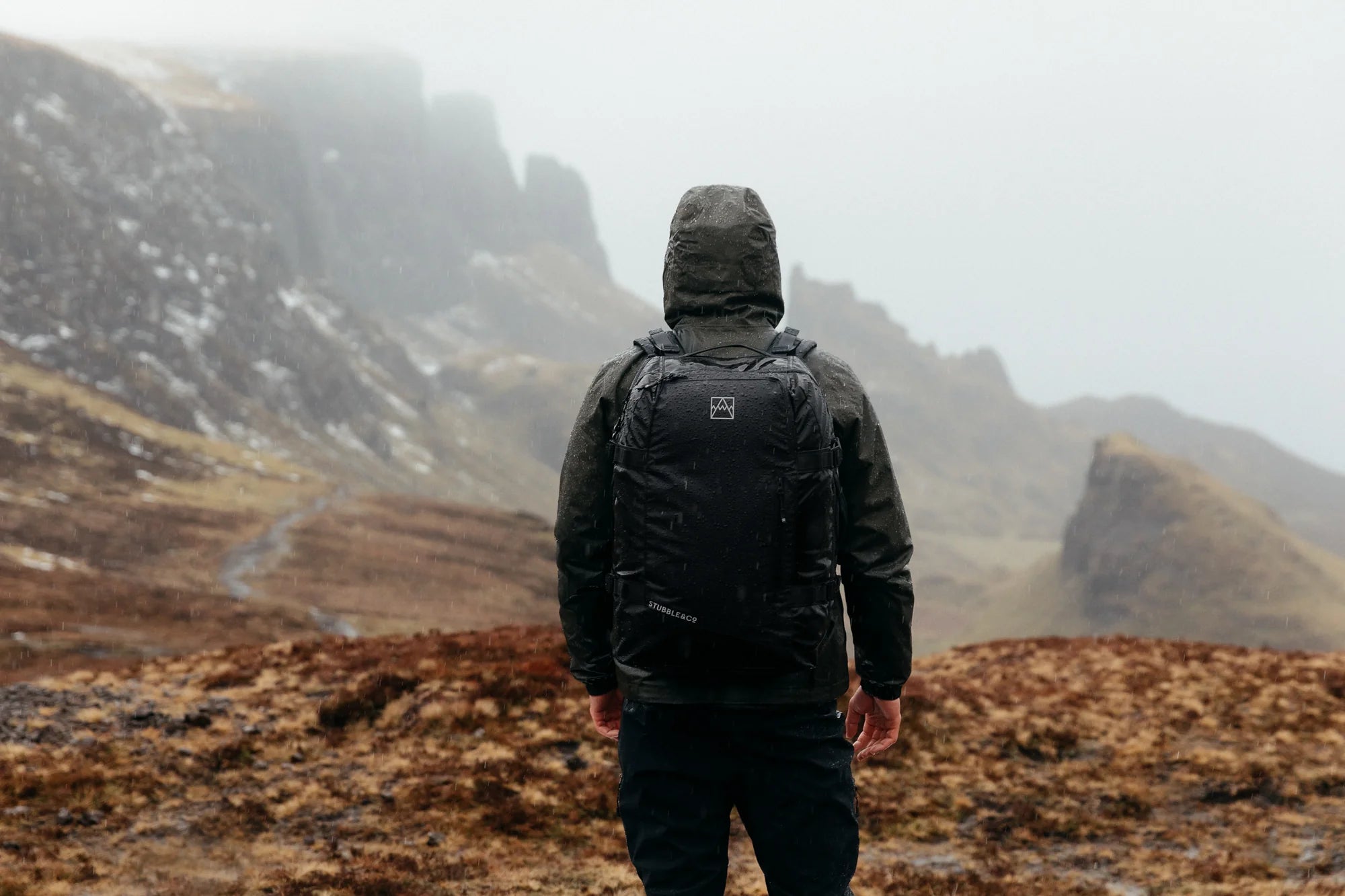 A man standing on a mountain in the rain wearing an Adventure Bag