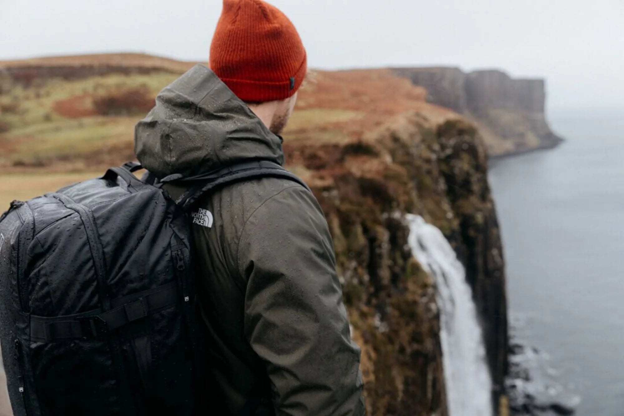 A man standing on a cliff edge by a waterfall wearing an Adventure Bag on his back