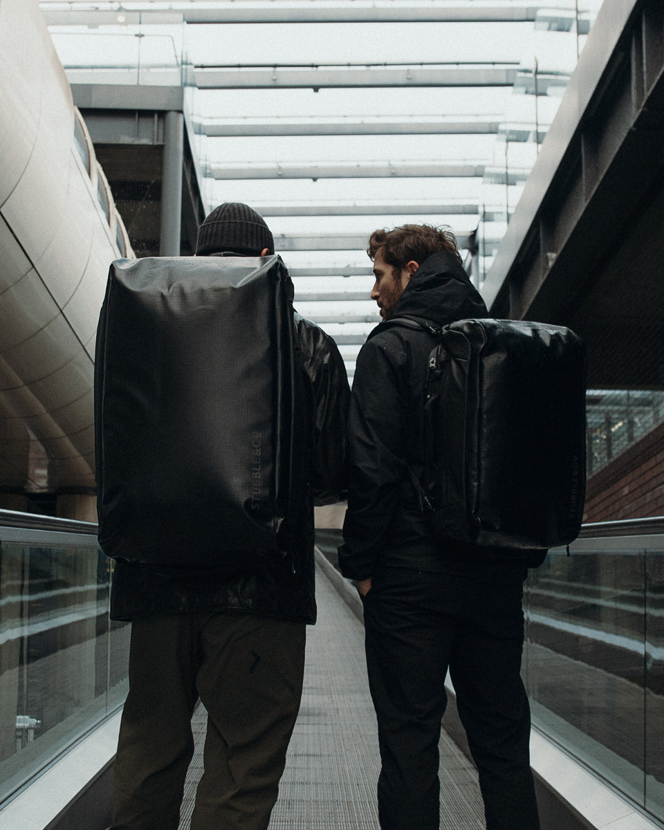 two men standing on an escalator wearing a kit bag 30l and a kit bag 65l