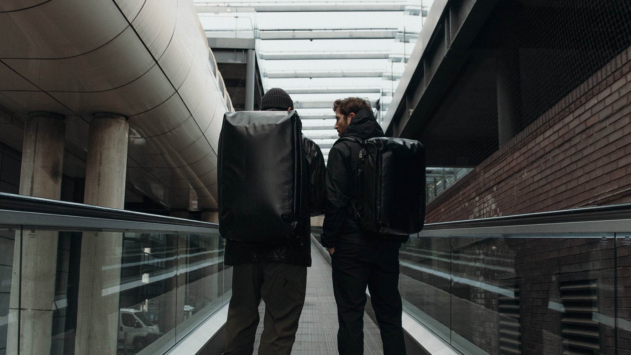 two men standing on an escalator wearing a 65L and 30L Kit bag on their backs