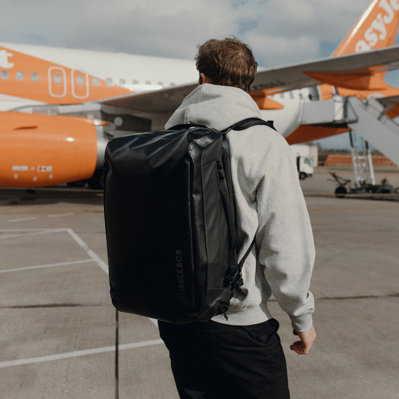 a man wearing an All Black Kit Bag 30L kit bag on his back walking towards an airplane