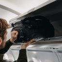 A man putting an All Black Kit Bag 30L in the overhead locker on an airplane