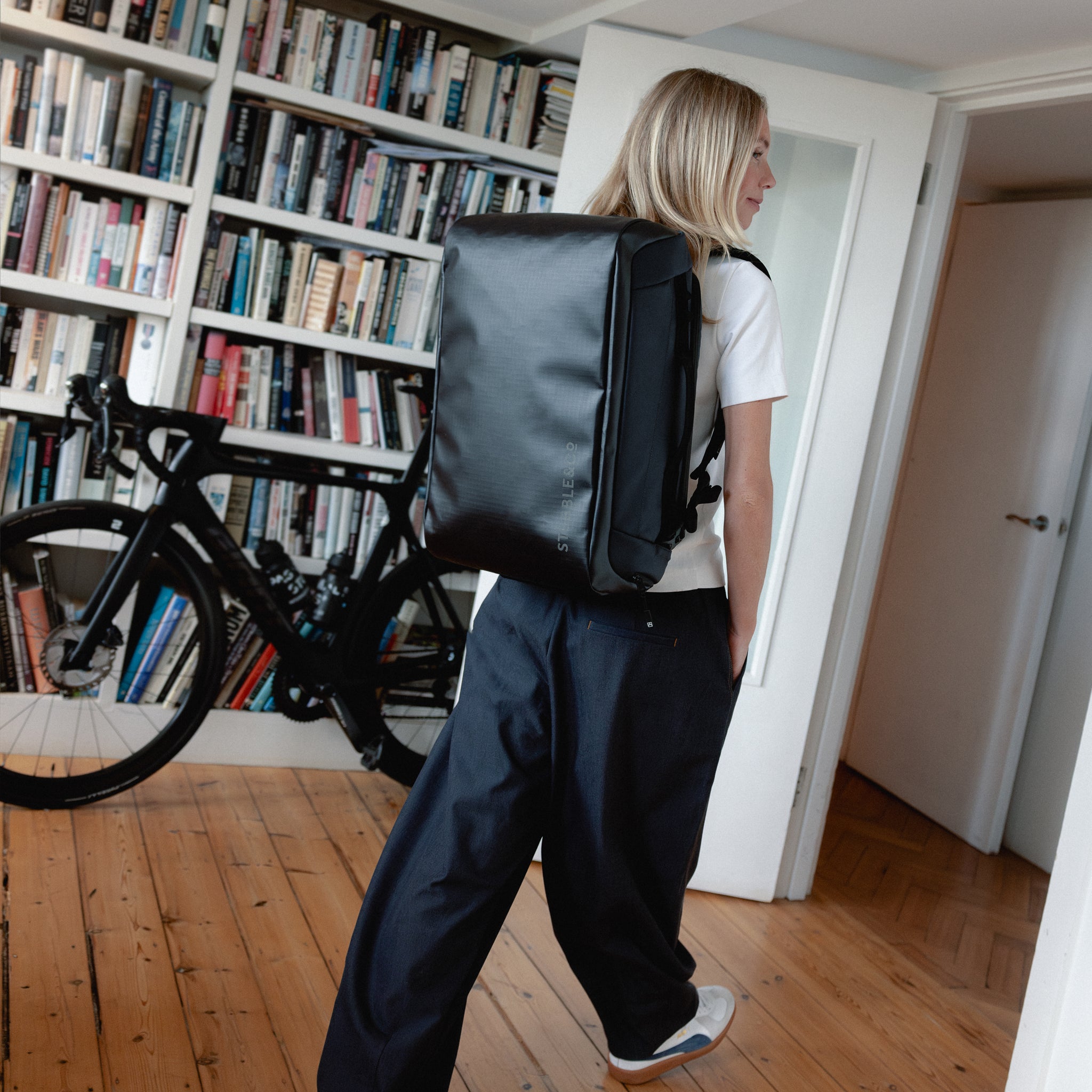 A man walking through the apartment wearing a Kit Bag 30L in Midnight Blue on her back
