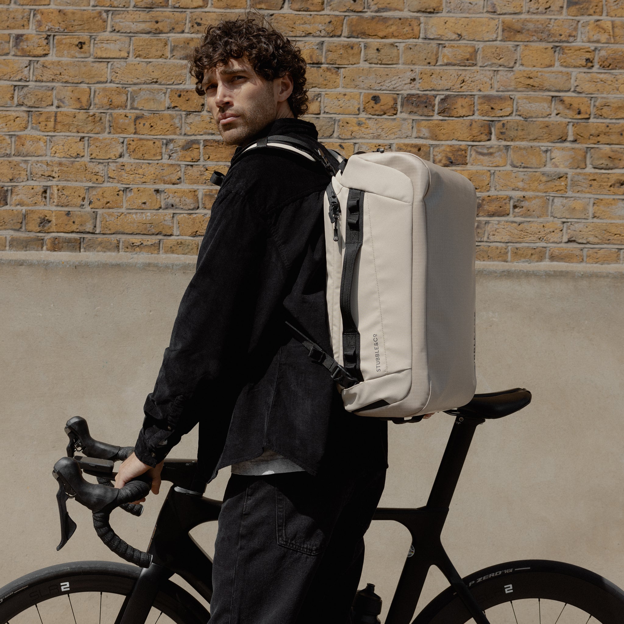 A man pushing a bike down the street wearing a Kit Bag 30L in Sand on his back