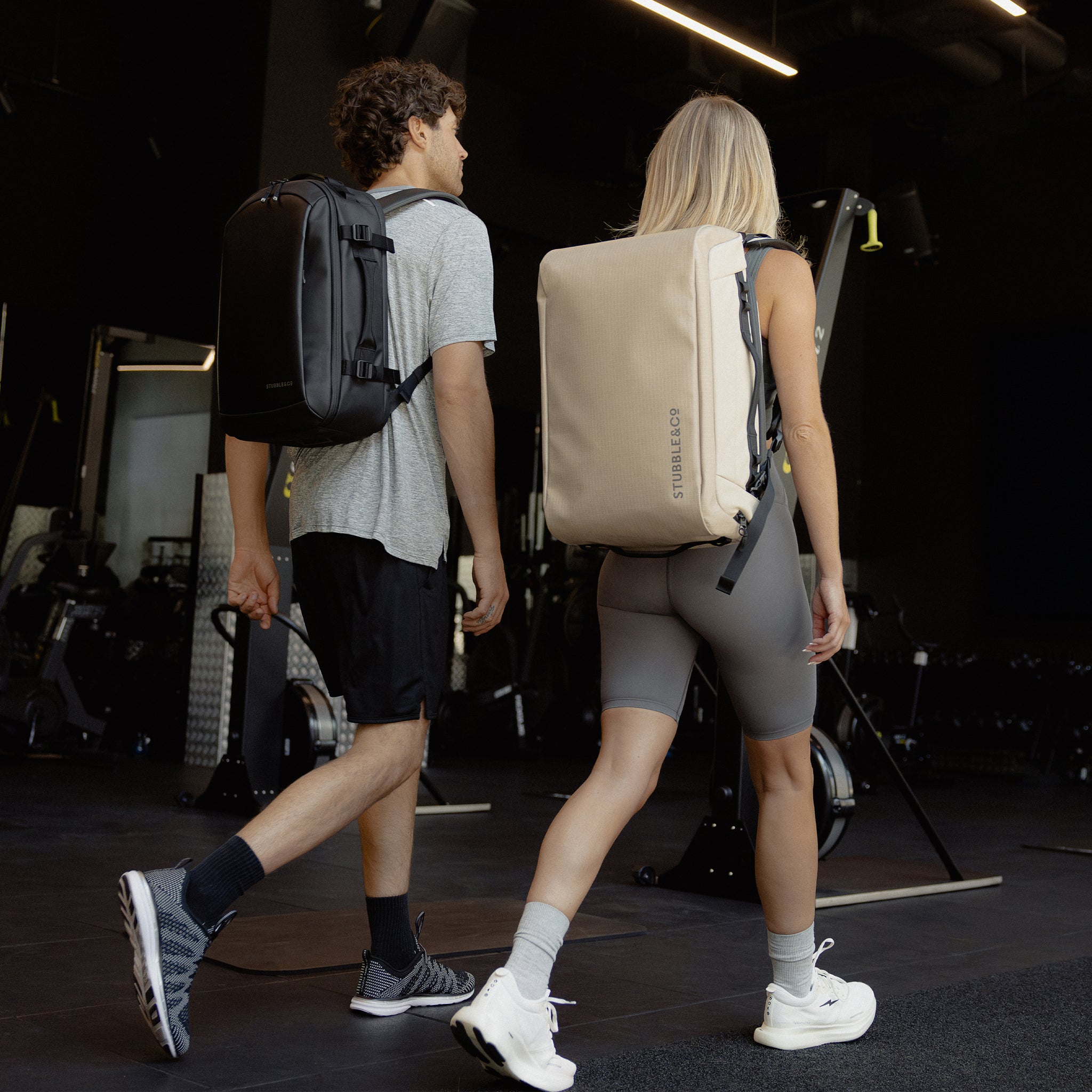 A man and woman wearing a backpacks on their backs as they walk through the gym including a Hybrid Backpack and Kit Bag 30L in Sand