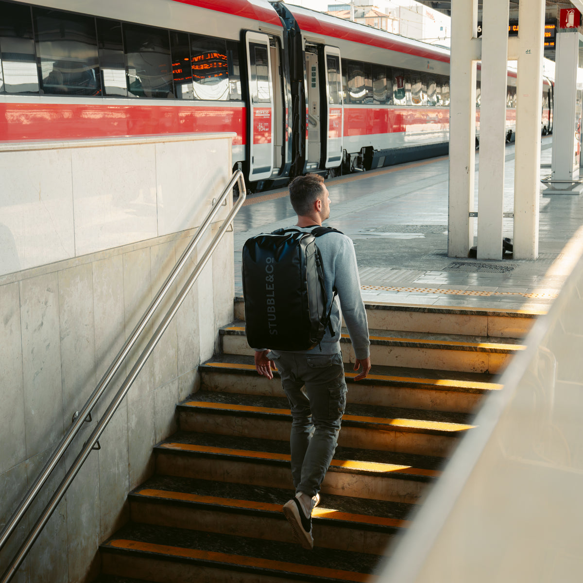 Man climbing station steps beside a train, wearing the Kit Bag 40L in All Black as a backpack