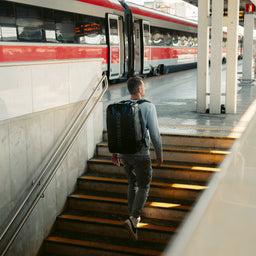 Man climbing station steps beside a train, wearing the Kit Bag 40L in All Black as a backpack