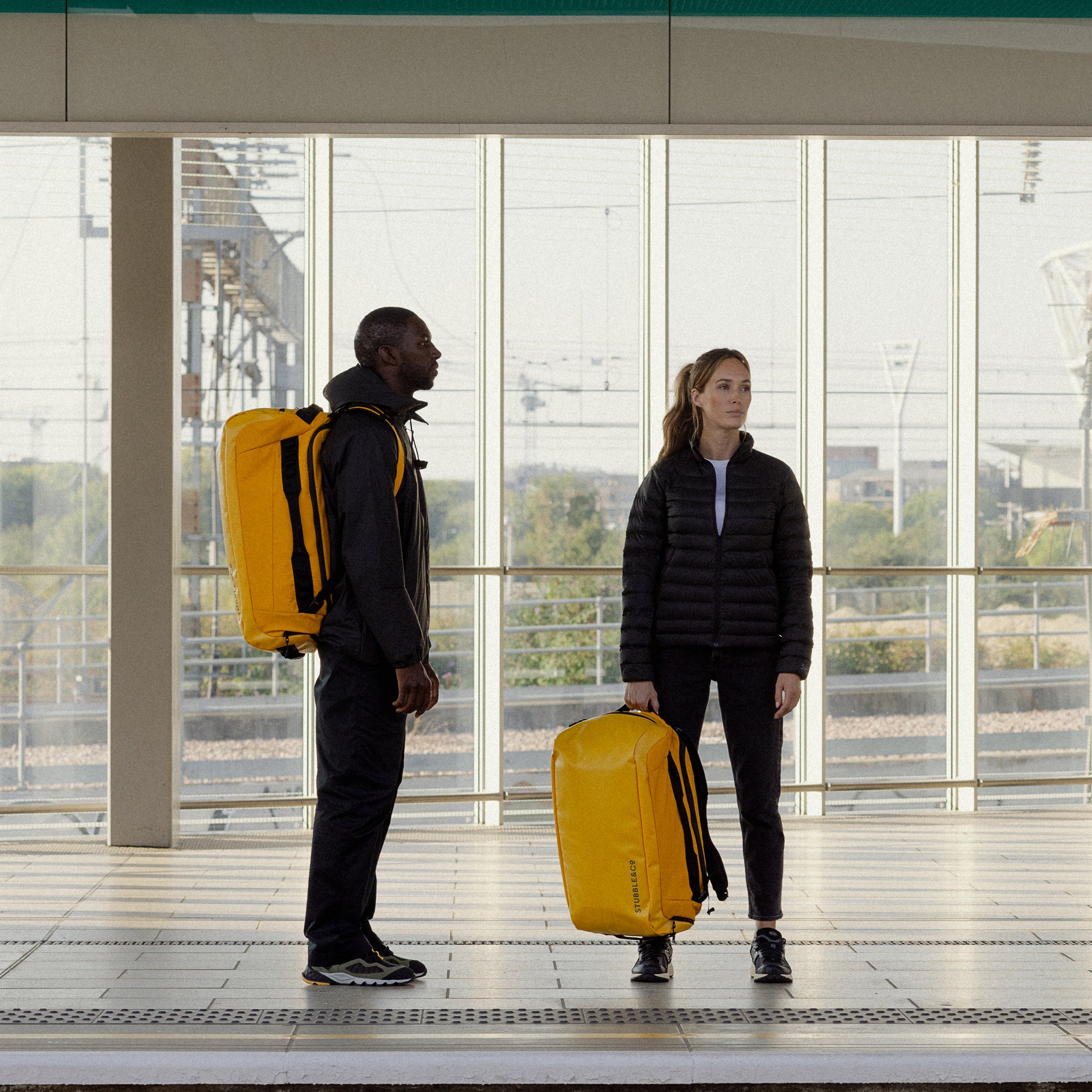 A man and woman standing on a train platform with Kit Bag 65L in Yellow