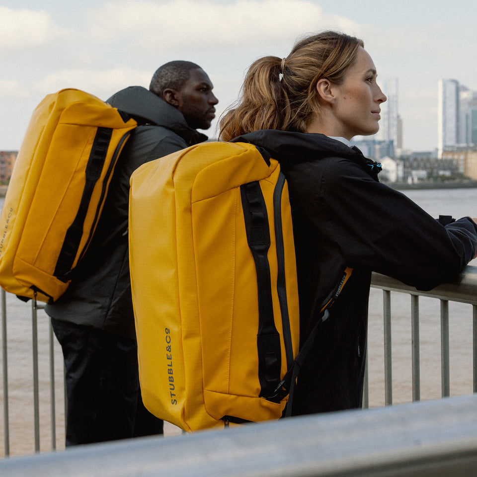 A man and woman standing next to eachother, both wearing a Kit Bag 65L in Yellow on their backs