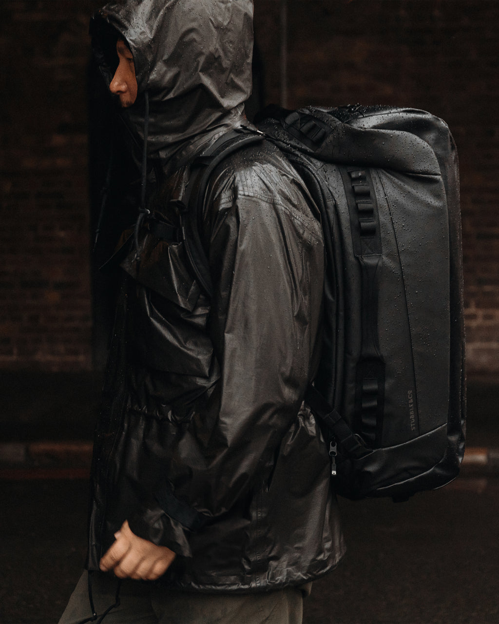 A man walking in front of a brick wall with his hood up and a kit bag 65l in black on his back in the rain