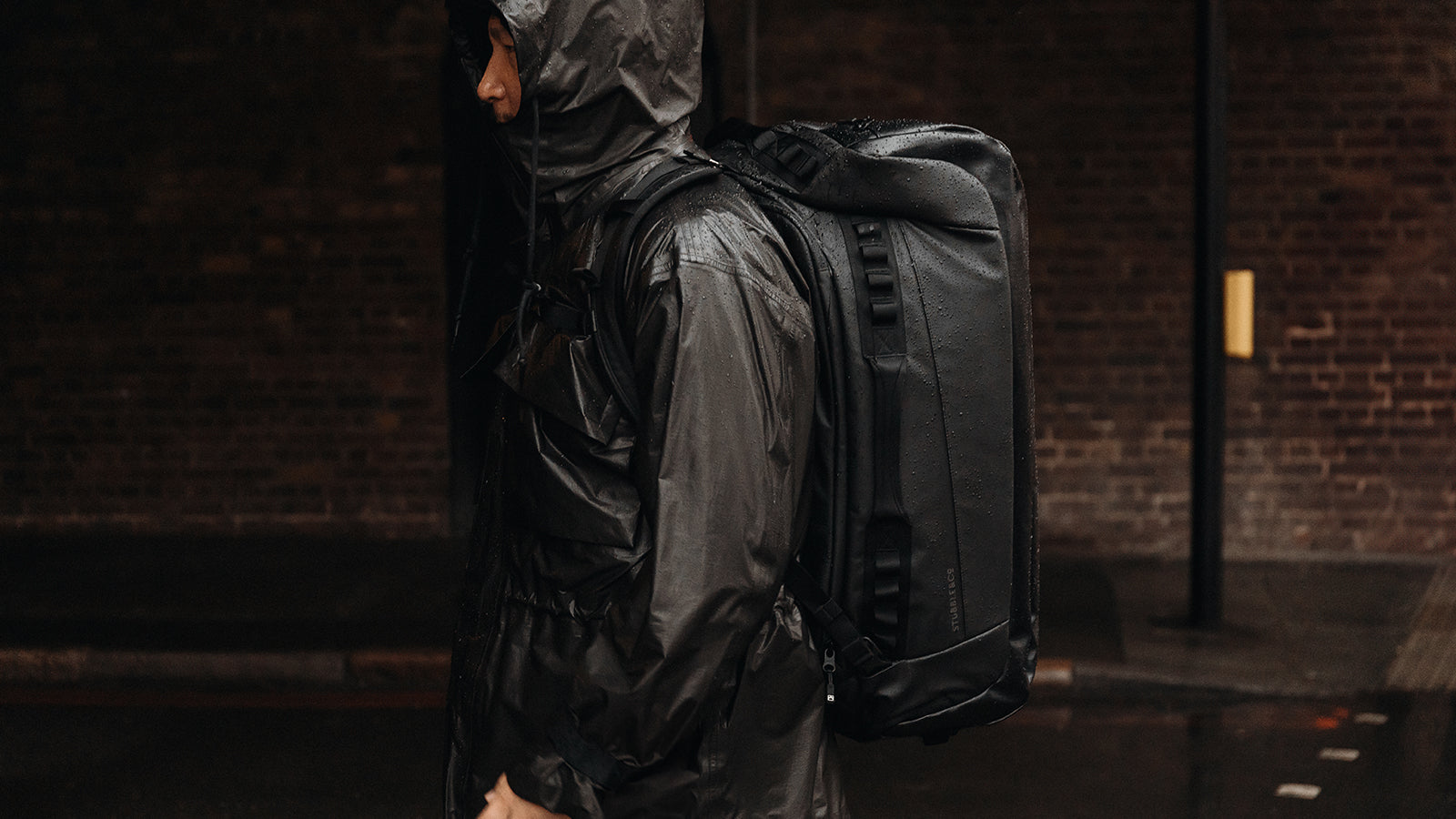 A man walking in front of a brick wall with his hood up and a kit bag 65l in black on his back in the rain- desktop