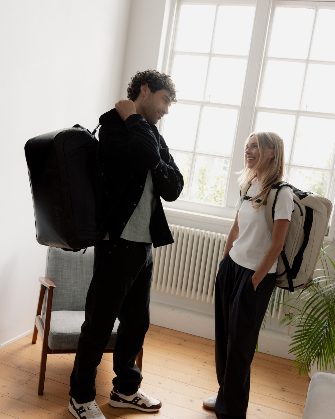 Man and woman standing in a London flat, wearing kit bags on their backs, his All Black and hers Sand.