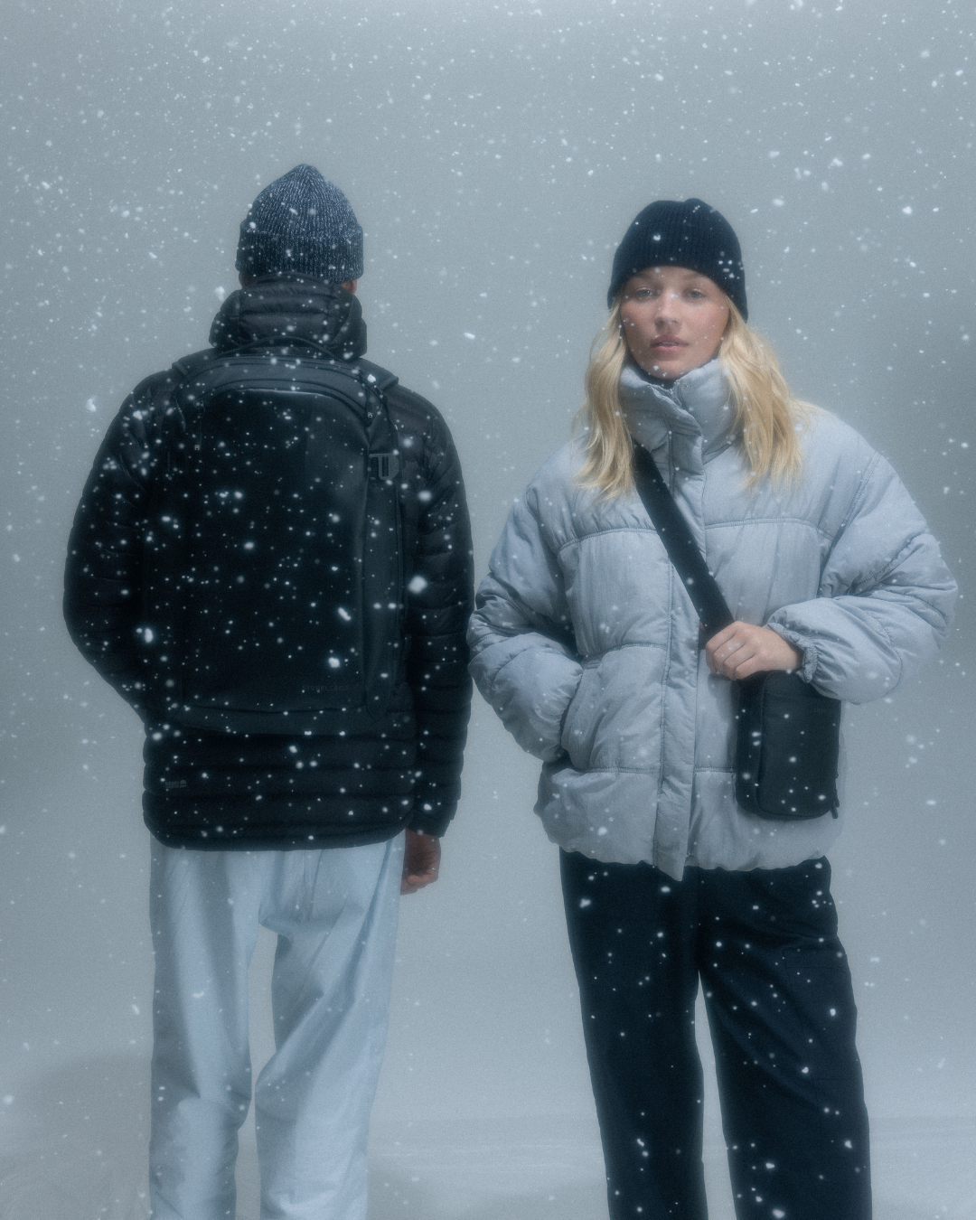 Man and woman wearing jackets and hats in the snow, the woman with a crossbody bag and the man with a backpack.
