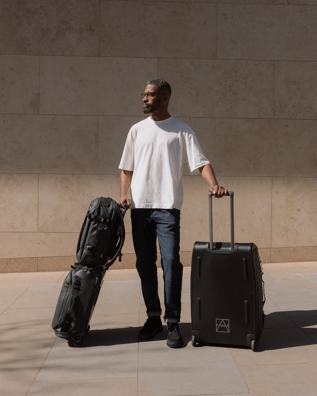 Man in a white T-shirt with the All Black Wheeled Duffel 80L, the All Black Wheeled Duffel 40L, and a backpack resting on top.