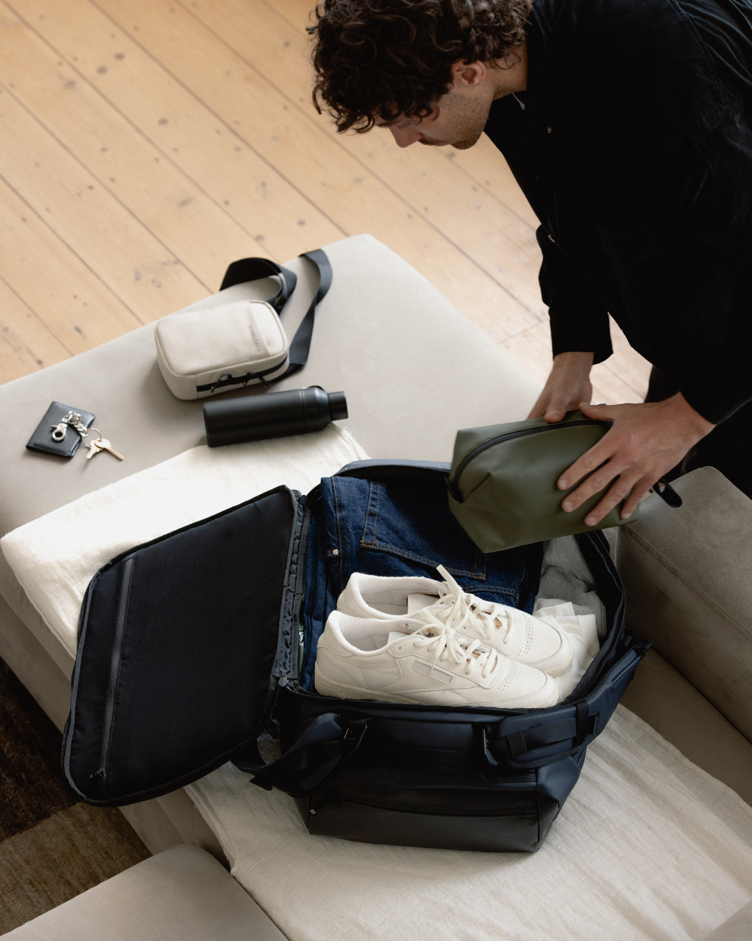 Man packing a kit bag with clothes, shoes and the Urban Green Wash Bag, with the Sand Shoulder Bag laid flat on the edge of a sofa.