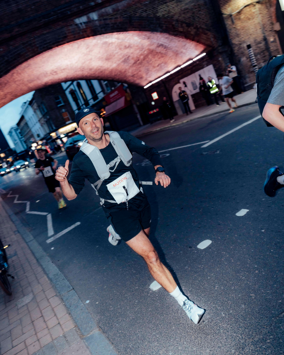 A man running under a bridge along the road with his thumbs up whilst wearing an ash Motion Backpack 20L on his back