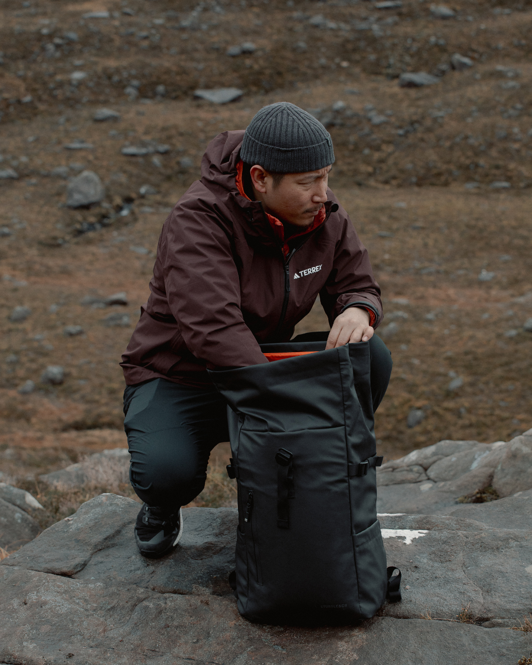Man standing in the countryside, rummaging through a roll top backpack with the orange interior partly visible.
