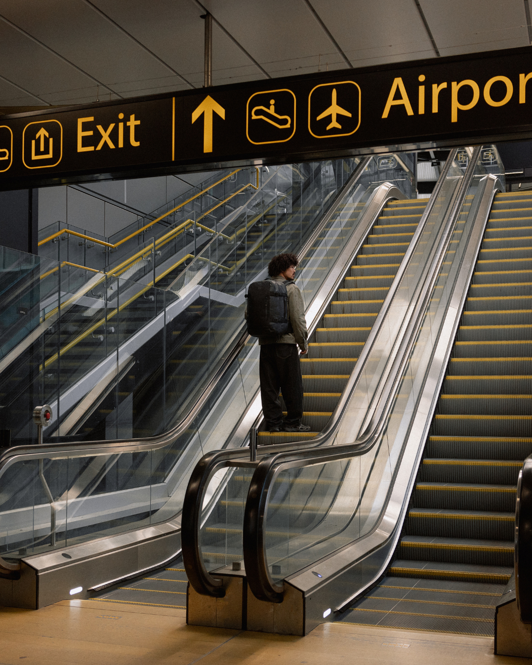 Man travelling up an escalator at the airport, wearing a black backpack.
