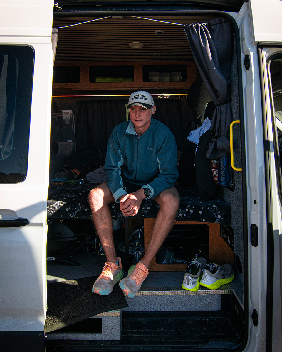 Man sits in the back of a white van wearing a blue fleece, white cap, and orange running shoes