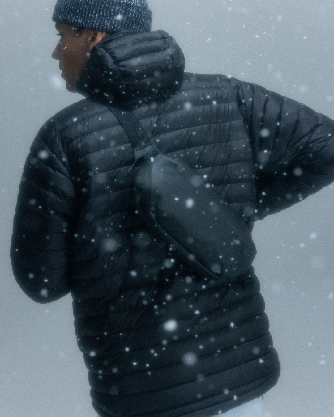 Man wearing a black crossbody bag and a black puffer jacket in the snow.