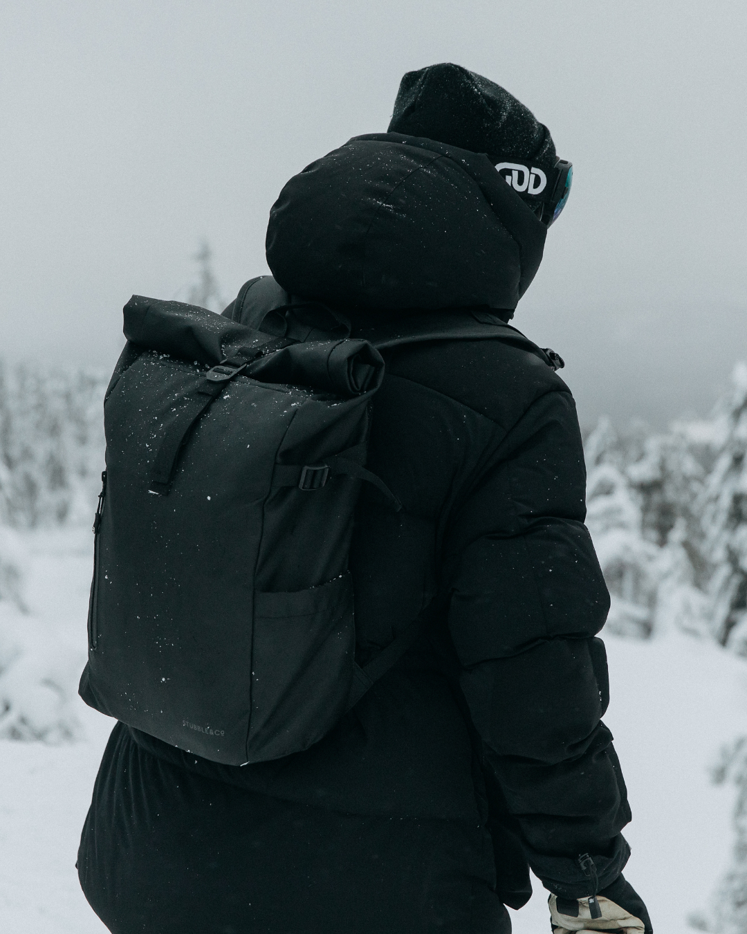 Man facing away from the camera, wearing the All Black Roll Top 20L backpack, with a snowy landscape ahead.