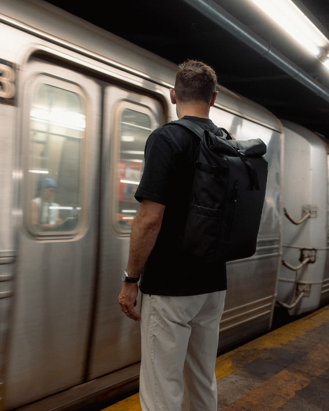 Man waiting for a train, wearing the All Black Roll Top Backpack.