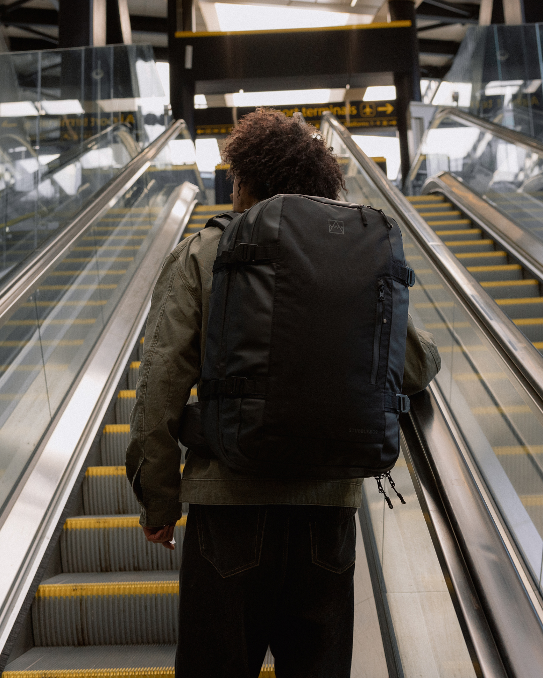 Man wearing the All Black Travel Backpack 40L, going up an airport escalator.