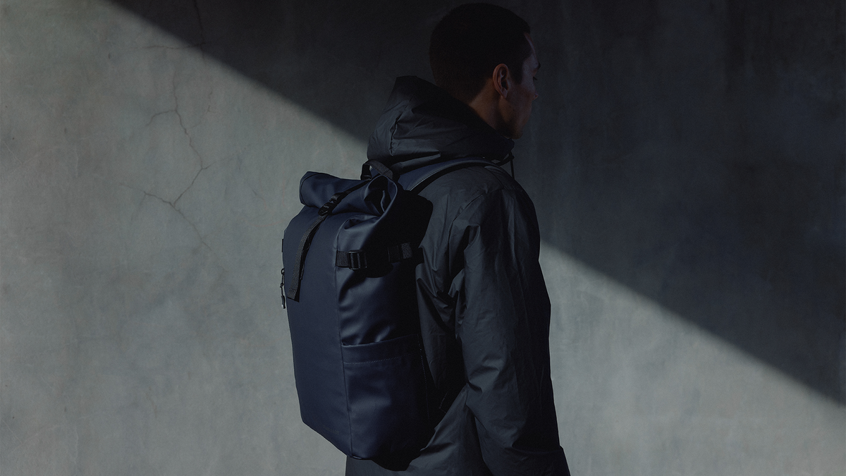 A man in a dark hooded jacket stands in profile wearing a dark navy waterproof backpack, set against a textured gray wall with dramatic diagonal light and shadow.